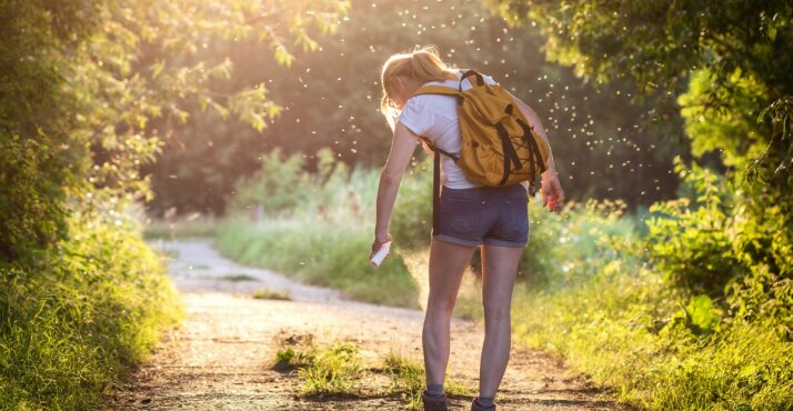 Eine Frau in der Natur, umgeben von Mücken. Mit einem Spray in der Hand.
